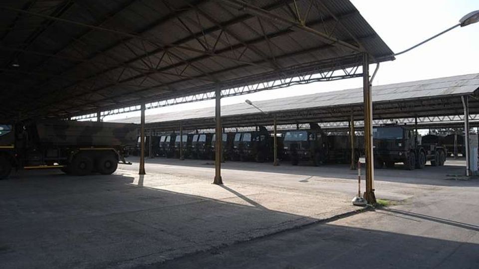 Ampia tettoia industriale con struttura metallica e copertura in lamiera, sostenuta da pilastri, che ombreggia un piazzale asfaltato. Sullo sfondo si vedono altre pensiline simili con diversi veicoli pesanti parcheggiati in fila.