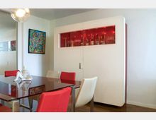 Sala da pranzo moderna con tavolo in vetro e sedie rosse e bianche, illuminata da una lampada a sospensione. Sullo sfondo un grande mobile bianco con vetrina superiore retroilluminata in rosso che espone bottiglie e bicchieri, con quadro colorato sulla parete.