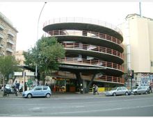 Edificio urbano a pianta circolare con più livelli sovrapposti e rampe elicoidali esterne, evidenziate da ringhiere rosse, affacciato su una strada trafficata. Alla base si vedono pilastri di sostegno, un’area d’ingresso e alberi e auto parcheggiate lungo il marciapiede.