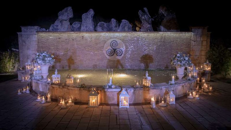 Fontana semicircolare in una piazza lastricata, illuminata di notte e circondata da numerose lanterne e candele. Sullo sfondo c’è un alto muro in pietra con un simbolo circolare e rocce decorative in sommità.