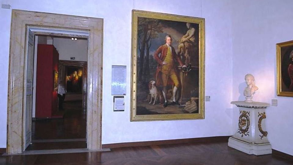 Sala di museo con pareti bianche e pavimento in parquet, con un grande dipinto incorniciato al centro e una scultura su piedistallo decorato sulla destra. A sinistra si vede un’ampia porta con cornice in pietra che conduce a un’altra galleria con pareti rosse e altre opere esposte.