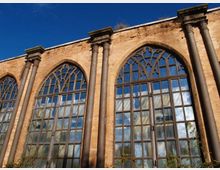 Facciata in mattoni di un edificio storico con alte finestre ad arco a sesto acuto e vetrate a riquadri con motivi gotici nella parte superiore. Pilastri verticali scuri scandiscono la parete sotto un cielo azzurro.