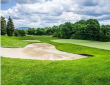 Campo da golf con green e bandierina, circondato da ampi prati e una grande buca di sabbia in primo piano. Sullo sfondo si vedono alberi fitti, colline lontane e un cielo nuvoloso.
