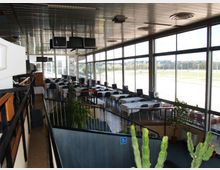 Interno di una sala ristorante o lounge con grandi vetrate a tutta altezza lungo un lato, tavoli apparecchiati e sedie disposte in file. Soffitto metallico con schermi sospesi, passerella con ringhiera e piante in vaso, con vista esterna su un’ampia area aperta.