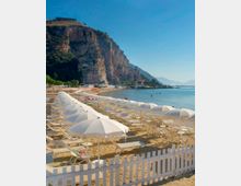 Spiaggia attrezzata con file di ombrelloni e lettini bianchi su sabbia dorata, delimitata da una staccionata bianca. Sullo sfondo si vede una grande falesia rocciosa che domina la costa e un mare calmo lungo la baia.