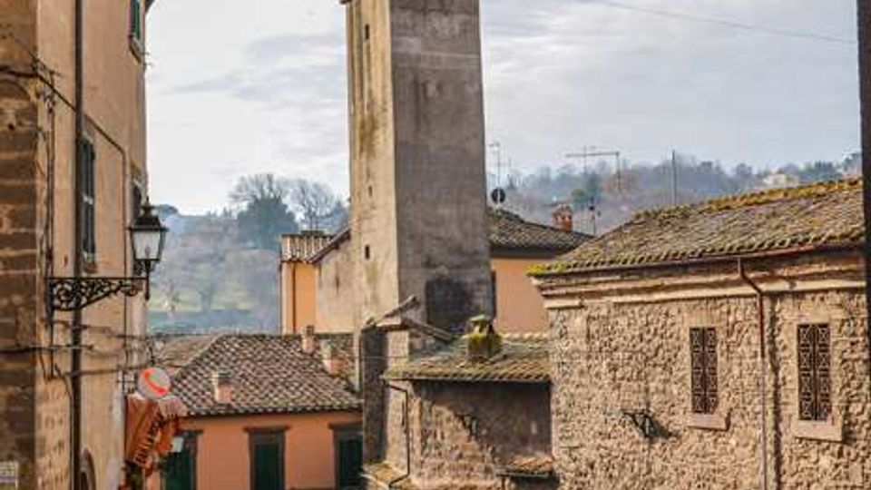 Strada in discesa in un borgo storico con case in pietra e intonaco, tetti in coppi e lampioni in ferro battuto. Al centro si vede una torre quadrata con struttura metallica in sommità, sullo sfondo colline e cielo nuvoloso.
