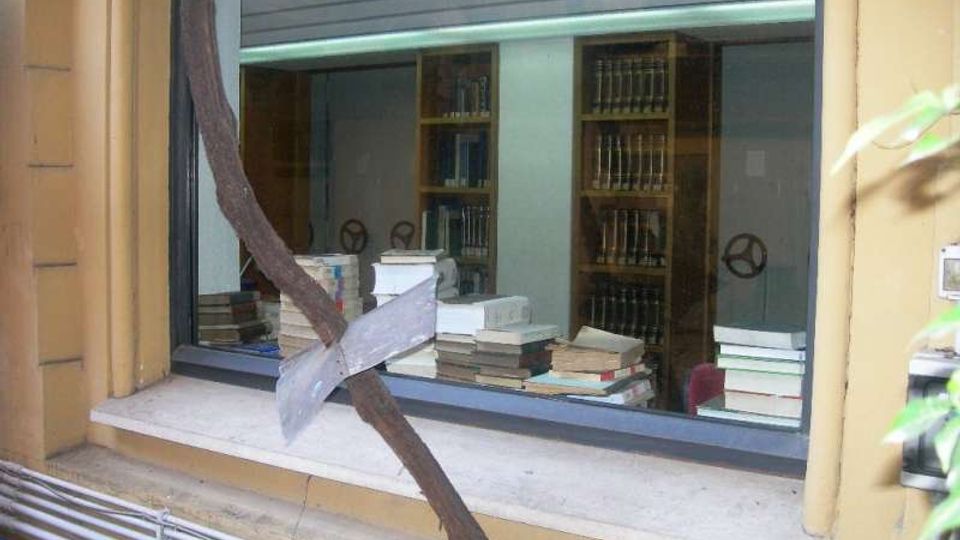 Finestra su strada che mostra l’interno di una biblioteca o archivio, con scaffali alti pieni di volumi e numerose pile di libri su un tavolo vicino al vetro. Davanti alla finestra passa una struttura metallica arrugginita e, sotto il davanzale, si vedono tubazioni esterne lungo la parete.