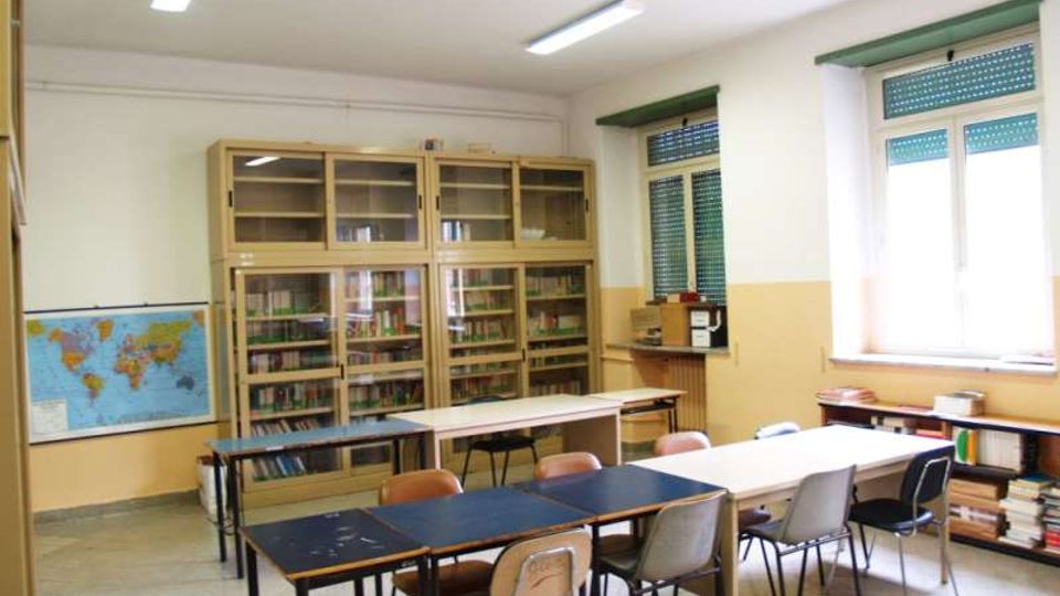 Aula scolastica con tavoli e sedie disposti in file, armadi vetrati pieni di libri lungo la parete e una carta geografica appesa. Grandi finestre con tapparelle verdi illuminano l’ambiente, con scaffali bassi e pile di libri vicino al davanzale.