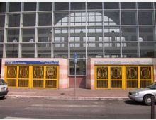 Ingresso di una stazione della metropolitana sotto una grande facciata vetrata a griglia, con due varchi affiancati chiusi da cancelli gialli e pannelli di segnaletica sopra le aperture. Davanti si vedono marciapiede e strada con auto parcheggiate.