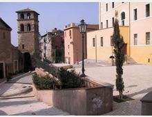 Piazza in pendenza di un borgo storico con pavimentazione in pietra, aiuole e lampioni in ferro battuto. Sullo sfondo si vedono una torre campanaria in pietra e facciate di edifici intonacati con finestre e scale esterne.