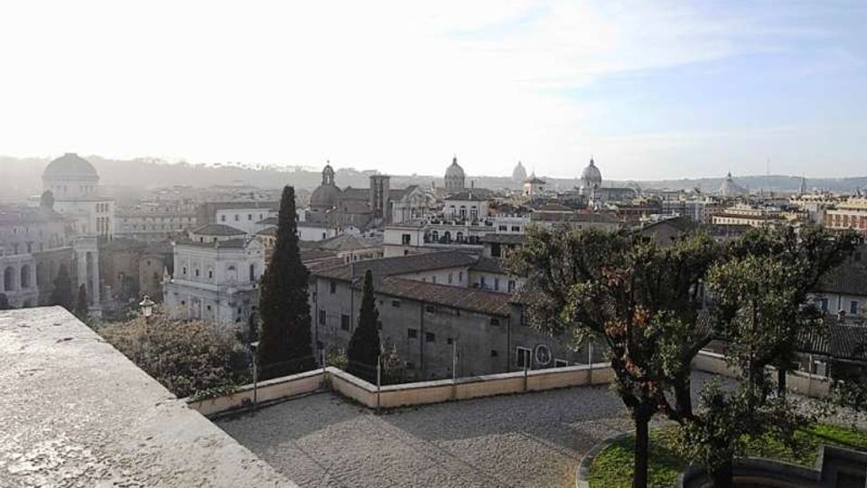 Veduta panoramica di una città storica con tetti in tegole e numerose cupole sullo sfondo, osservata da una terrazza in pietra. In primo piano si vedono una piazzetta lastricata, un muretto perimetrale e alberi lungo il bordo del belvedere.