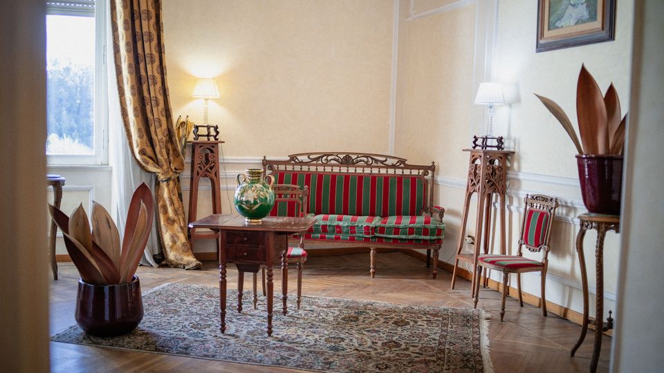 Salotto in stile classico con pavimento in parquet e tappeto, divano e sedie imbottite a righe rosse e verdi, tavolino in legno e lampade su alti piedistalli intagliati. Una grande finestra con tende pesanti illumina la stanza, con vasi decorativi e un quadro appeso alla parete.