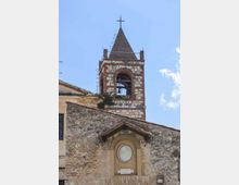 Campanile di una chiesa in muratura mista di pietra e mattoni, con aperture ad arco e campane visibili, sormontato da una guglia con croce. In primo piano si vede il tetto in tegole e una nicchia con tondo decorativo sulla facciata, sotto un cielo azzurro.