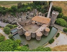 Veduta aerea di un castello in pietra circondato da un fossato d’acqua, con mura e torri cilindriche e un edificio centrale con tetto in tegole. Intorno si vedono un ponte d’accesso, una strada e aree verdi e campi coltivati ai margini.