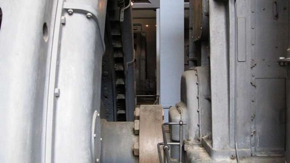 Interno di un impianto industriale o museo tecnico con grandi macchinari metallici grigi affiancati, tubazioni e componenti meccanici visibili. In primo piano si nota una ringhiera di sicurezza lungo un passaggio stretto tra le strutture.