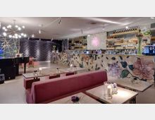 Interno di un bar-lounge moderno con divanetti rosa e tavolini in marmo illuminati da candele, e lungo bancone decorato con motivi floreali e scaffali di bottiglie sullo sfondo. A sinistra si vede un pianoforte a coda e lampadari sferici, con pareti scure a texture ondulata e illuminazione a faretti.