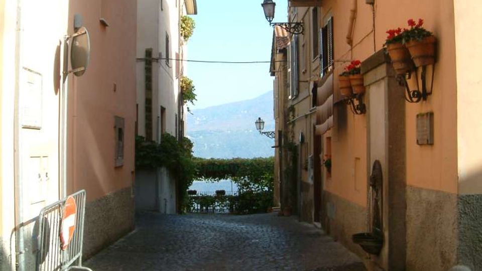 Vicolo stretto in ciottoli tra edifici dai colori chiari, con lampioni a parete e balconi con vasi di fiori. In fondo alla strada si intravede un lago con montagne sullo sfondo.