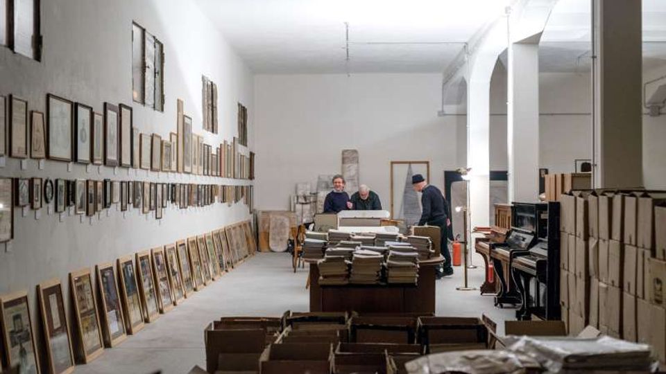 Ampio locale interno tipo deposito o archivio con pareti bianche e soffitto alto, con molti quadri e cornici appoggiati a terra e appesi lungo la parete sinistra. Al centro e in primo piano si vedono tavoli e scatole con pile di documenti e libri, mentre a destra ci sono colonne ad arco e alcuni pianoforti vicino a scaffalature.