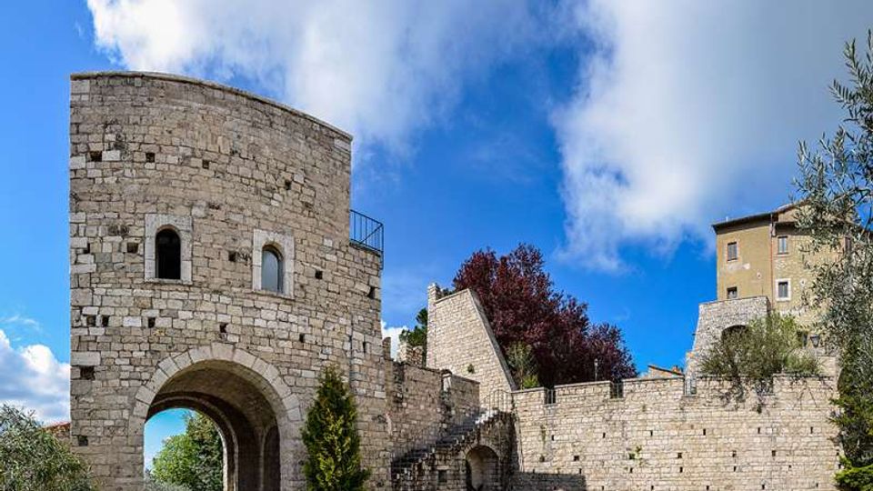 Porta fortificata in pietra con torre semicircolare e grande arco d’ingresso, collegata a mura merlate con una scala laterale. Sullo sfondo si vedono alberi e un edificio in pietra sotto un cielo azzurro con nuvole.
