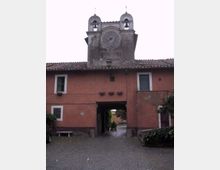 Facciata di un edificio storico intonacato color rosso con tetto in coppi e passaggio ad arco centrale che conduce a un cortile interno. Sopra l’ingresso si alza una torre con grande orologio e due campanili gemelli con campane, sormontati da croci.