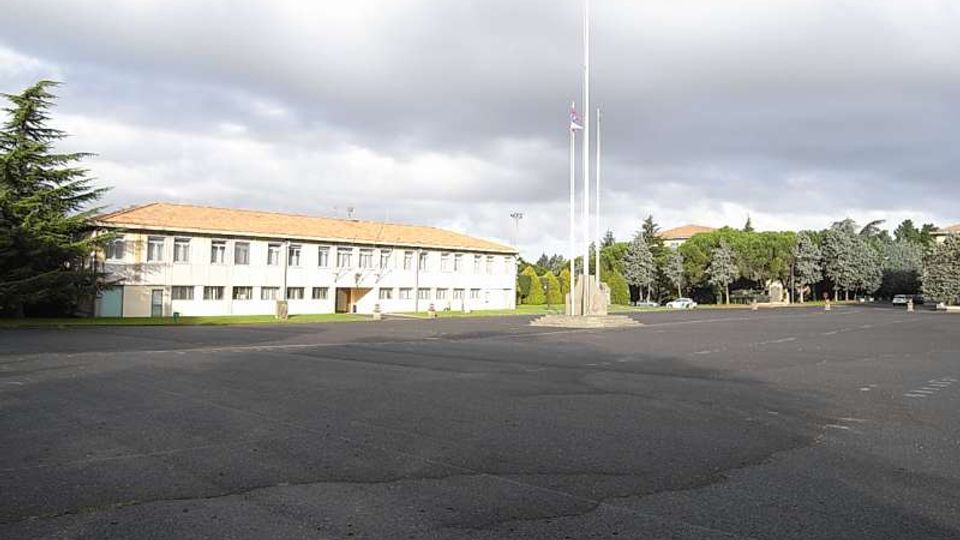 Ampio piazzale asfaltato con al centro alti pennoni portabandiera, davanti a un edificio basso e allungato a due piani con tetto in coppi e facciata chiara. Sullo sfondo si vedono alberi e altre costruzioni, sotto un cielo nuvoloso.