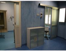 Interno di una stanza clinica con pareti e pavimento blu, porte scorrevoli con pannelli in vetro e una finestra luminosa. Sono presenti un armadietto basso, attrezzature a parete e un’area adiacente con carrello metallico e postazione con sgabello.