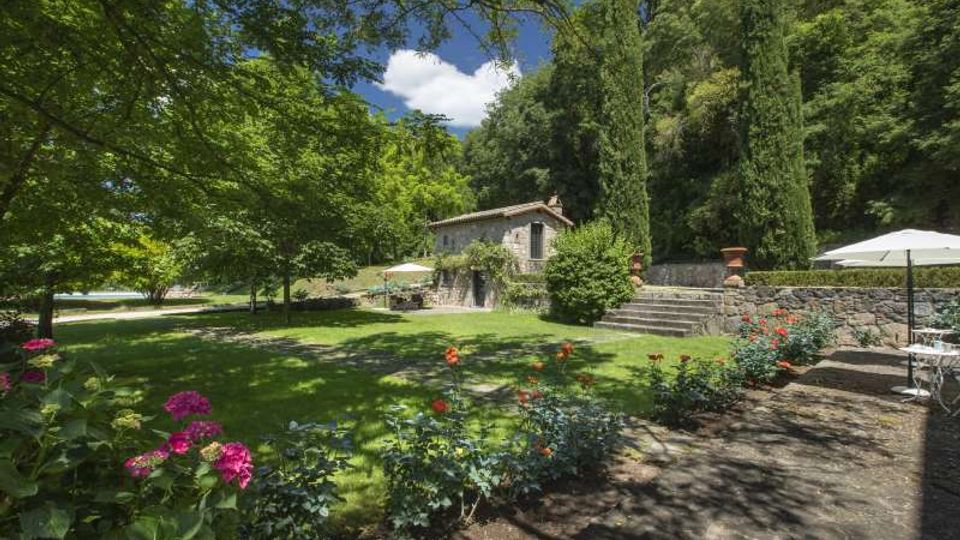 Giardino paesaggistico con prato ampio, aiuole fiorite e vialetto in pietra con tavolini all’aperto, ai piedi di un pendio boscoso. Sullo sfondo si vede una casa in pietra con tetto in coppi, una scalinata e cipressi accanto a un muretto di contenimento.
