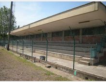 Tribuna di un impianto sportivo all’aperto con gradinate in cemento sotto una lunga copertura piatta, vista dietro una recinzione metallica. Nell’area circostante si notano un palo alto e alberi sullo sfondo.