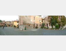 Piazza lastricata in pietra con edifici storici in muratura dai toni rosati e ocra, finestre con persiane e piccoli archi alla base. Ai bordi si vedono vasi con piante e un edificio parzialmente coperto da vegetazione, con ingressi e dettagli architettonici tradizionali.