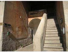 Cortile interno con pareti in intonaco consumato e un’ampia scala in pietra che sale a destra, affiancata da un alto parapetto. Sul fondo si vede un arco che conduce a un passaggio coperto e a un ingresso.