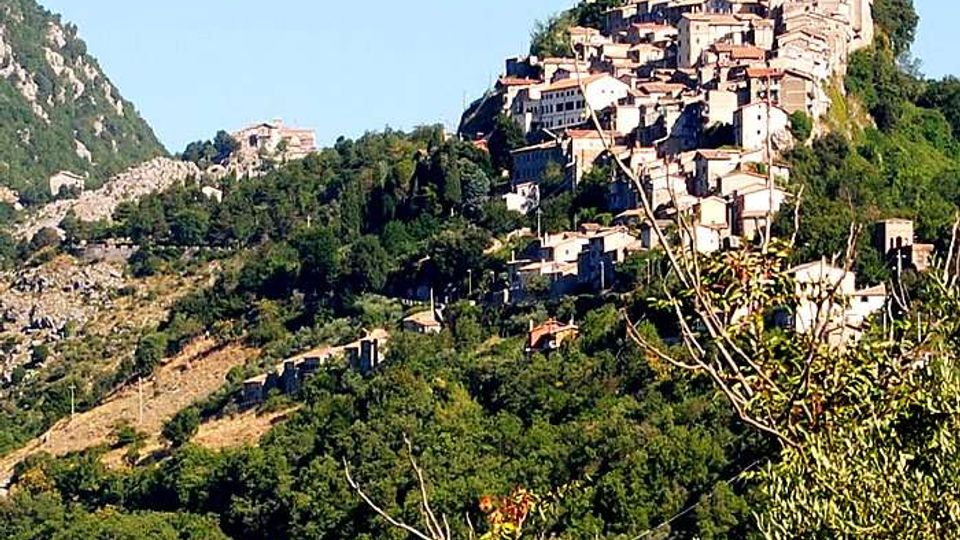 Borgo collinare con case in pietra addossate tra loro, arroccato su un pendio boscoso. Intorno si vedono colline verdi e un paesaggio montano sotto cielo sereno.