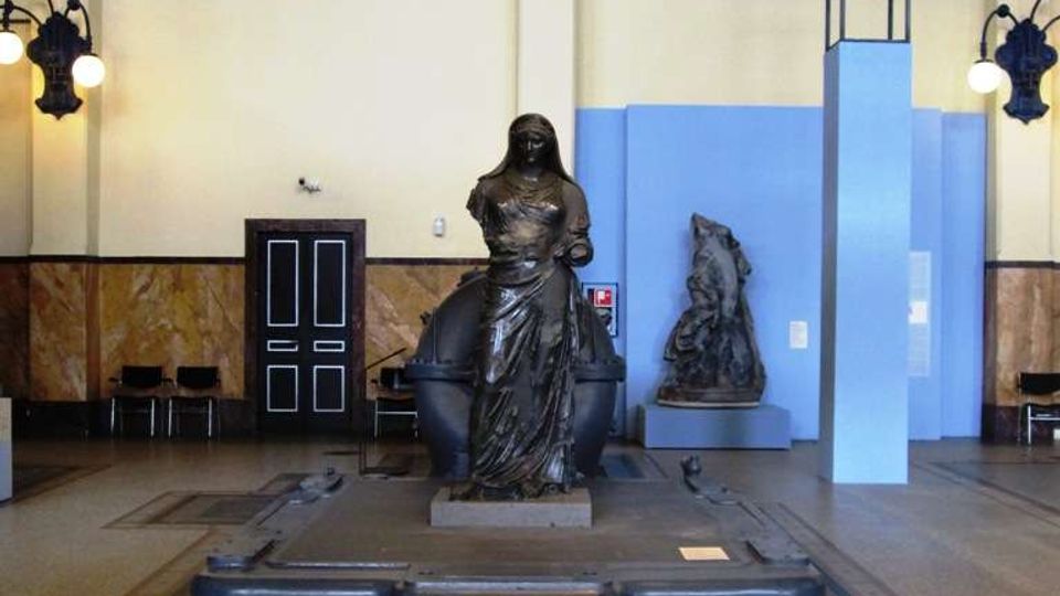 Sala interna in stile industriale con pavimento a mosaico e pareti chiare con pannelli blu; al centro una grande scultura in bronzo su basamento, circondata da una struttura metallica incassata nel pavimento. Sullo sfondo si vedono una porta, sedute laterali, lampade a parete e un secondo rilievo esposto su piedistallo.