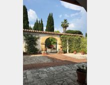 Cortile pavimentato in pietra con una parete intonacata e tetto in coppi, parzialmente coperta da edera, con un arco centrale che incornicia un cancello in ferro battuto. Sullo sfondo si vedono alti cipressi e una palma sotto un cielo azzurro con nuvole.