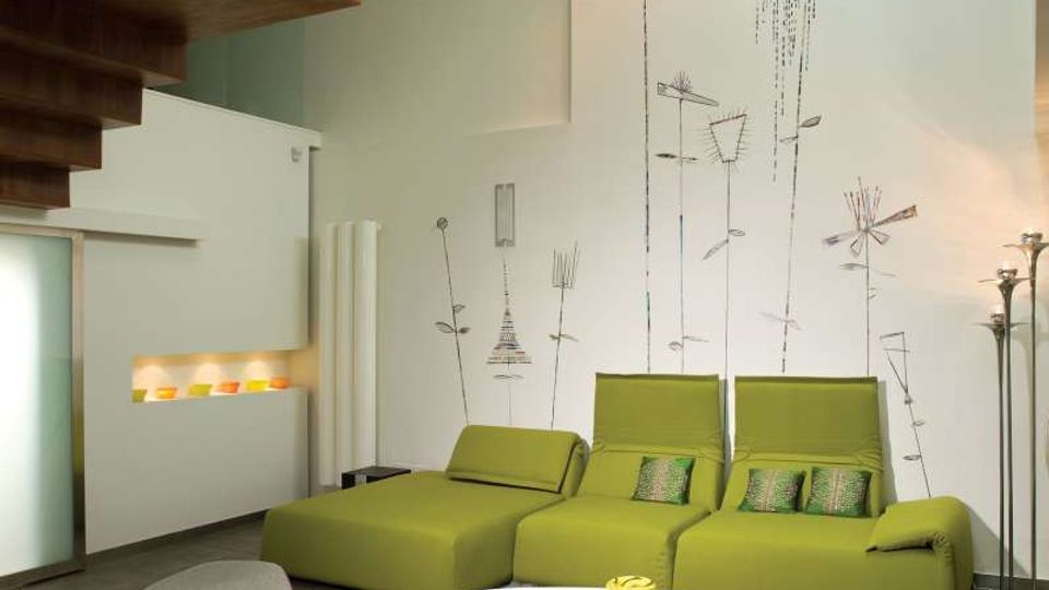 Soggiorno moderno con due divani chaise longue verdi e tavolino basso bianco su pavimento grigio. Parete bianca con decorazioni grafiche stilizzate, scala in legno a vista e nicchia illuminata con oggetti colorati.