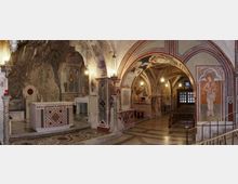 Interno di una chiesa o cappella sotterranea con navate ad archi e volte affrescate, pareti in parte scavate nella roccia e pavimento a mosaico. In primo piano si vede un altare in pietra e, sul fondo, una porta finestrata con panche lungo il corridoio.