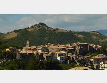 Veduta panoramica di un borgo collinare con case ravvicinate dai tetti in tegole e un campanile che svetta tra gli edifici. Sullo sfondo si vedono colline boscose e pendii verdi sotto un cielo parzialmente nuvoloso.
