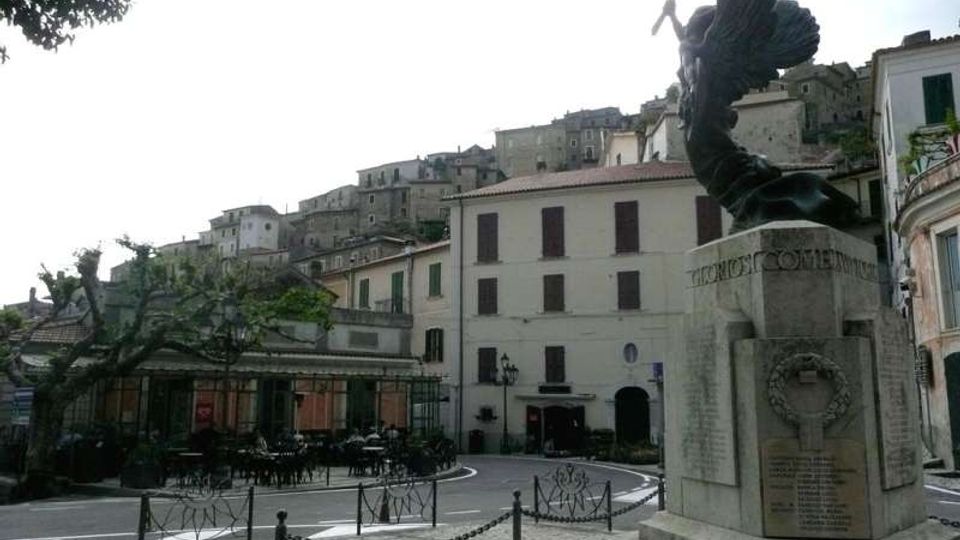Piazza di un borgo storico con pavimentazione in pietra e un monumento in marmo in primo piano, delimitato da catenelle. Sullo sfondo si vedono edifici in muratura con persiane e case addossate su un pendio, oltre a un’area con tavolini all’aperto sotto una struttura leggera.