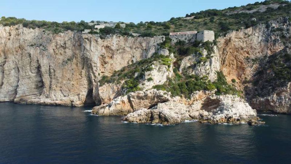 Scogliera costiera di roccia chiara a picco sul mare, con un promontorio roccioso coperto di macchia mediterranea. In alto sul crinale si vede una struttura in pietra simile a un forte o edificio storico, circondata da vegetazione.