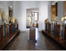Sala museale con pareti bianche e pavimento in pietra, con una statua classica al centro e busti in marmo esposti su supporti in legno e metallo lungo entrambi i lati. Sul fondo si vede un’apertura verso un’altra stanza con una finestra e una porta in legno.