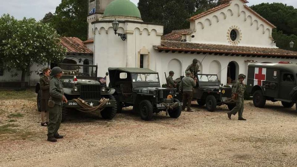 Piazzale sterrato davanti a una chiesa bianca con tetto in coppi, facciata ad archi e campanile con cupola verde e orologio. Nell’area sono parcheggiati diversi veicoli militari d’epoca, tra cui un mezzo con croce rossa, con alberi sullo sfondo sotto cielo nuvoloso.