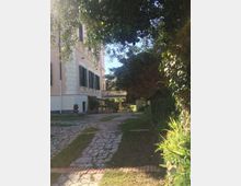 Vialetto in pietra e mattoni che attraversa un giardino alberato accanto a una villa in stile classico con facciata rosa e persiane verdi. Vegetazione fitta e alberi ombreggiano il percorso, con una balaustra e terrazza visibili sullo sfondo.