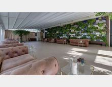 Ampio salone coperto con soffitto bianco a pannelli e pavimento chiaro, arredato con divani capitonné color cipria e tavolini in vetro con candele. Sul fondo si vede una grande parete verde verticale con piante e pannelli decorativi, affiancata da tende e un albero ornamentale.