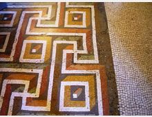 Pavimento a mosaico con motivi geometrici intrecciati e riquadri colorati in ocra, rosso e verde, delineati da tessere bianche. Sul lato destro si vede una fascia di pavimentazione in piccoli cubetti di pietra chiara.
