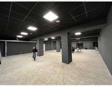 Ampio locale interno tipo magazzino o open space industriale con pareti grigio scuro, pavimento in cemento e colonne rettangolari. Soffitto a griglia nera con pannelli luminosi a LED e alcune attrezzature e tavoli sullo sfondo.