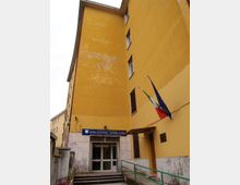 Ingresso di un edificio scolastico a più piani con facciata gialla, porta vetrata e insegna blu “Liceo Statale Ilaria Alpi”. Accanto all’entrata sono visibili una rampa con corrimano verde e due bandiere (Italia e Unione Europea) fissate al muro.