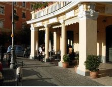 Portico curvo di un edificio in stile classico con grandi colonne chiare e decorazioni a rilievo, affacciato su un marciapiede in pietra. Sotto il portico si trovano tavolini e sedie di un bar con ombrelloni chiusi e vasi con piante, accanto a una strada con auto parcheggiate e lampione.
