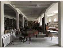 Ampia sala interna tipo archivio o laboratorio, con colonne bianche che sostengono il soffitto e scaffalature piene di libri e materiali lungo le pareti. Al centro ci sono un pianoforte a coda e tavoli da lavoro, con finestre sulla destra che illuminano l’ambiente.