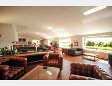 Ampia sala lounge interna con divani e poltrone in pelle trapuntata disposti in più zone, tavolini bassi e pavimento piastrellato. Grandi finestre laterali illuminano l’ambiente e affacciano su un’area verde esterna, con soffitto chiaro e luci a pannello.