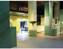 Interno di un museo o spazio espositivo con grandi pilastri e pareti verde chiaro, pavimento in piastrelle e illuminazione a soffitto. Su piedistalli sono esposte sculture e un grande bacile in pietra, con una passerella e un soppalco visibili sullo sfondo.