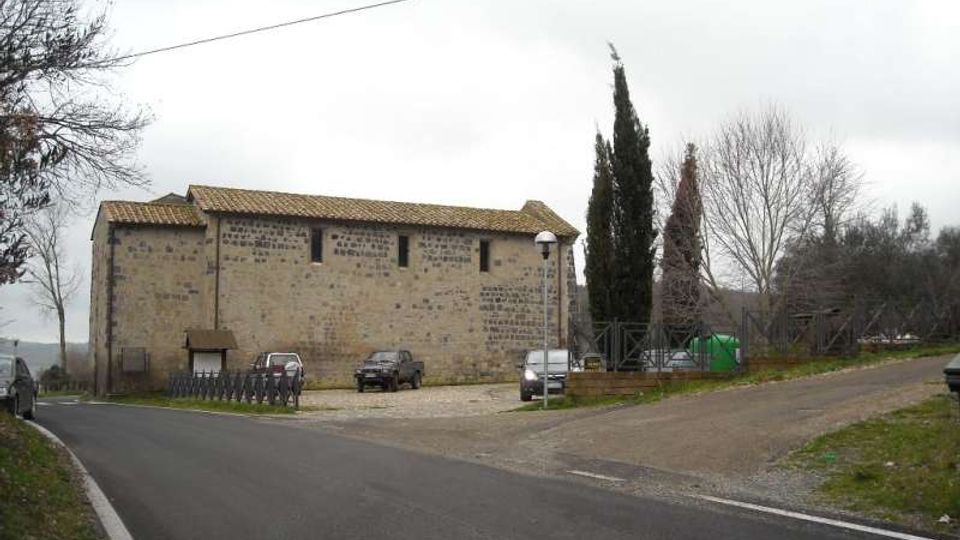 Edificio rurale in pietra con tetto a falde in tegole, affacciato su una strada asfaltata che curva e si biforca. Davanti si vede un piccolo parcheggio sterrato con recinzione, lampioni e alberi alti e spogli sullo sfondo.